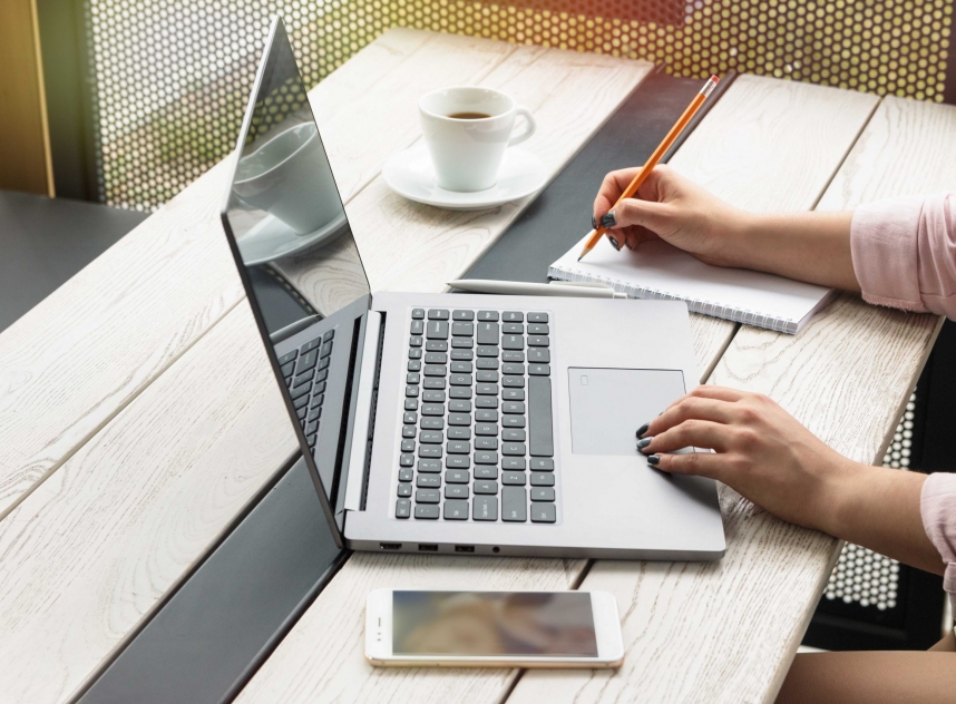 Close up portrait of a young woman working on laptop and writing, coffee on table.
